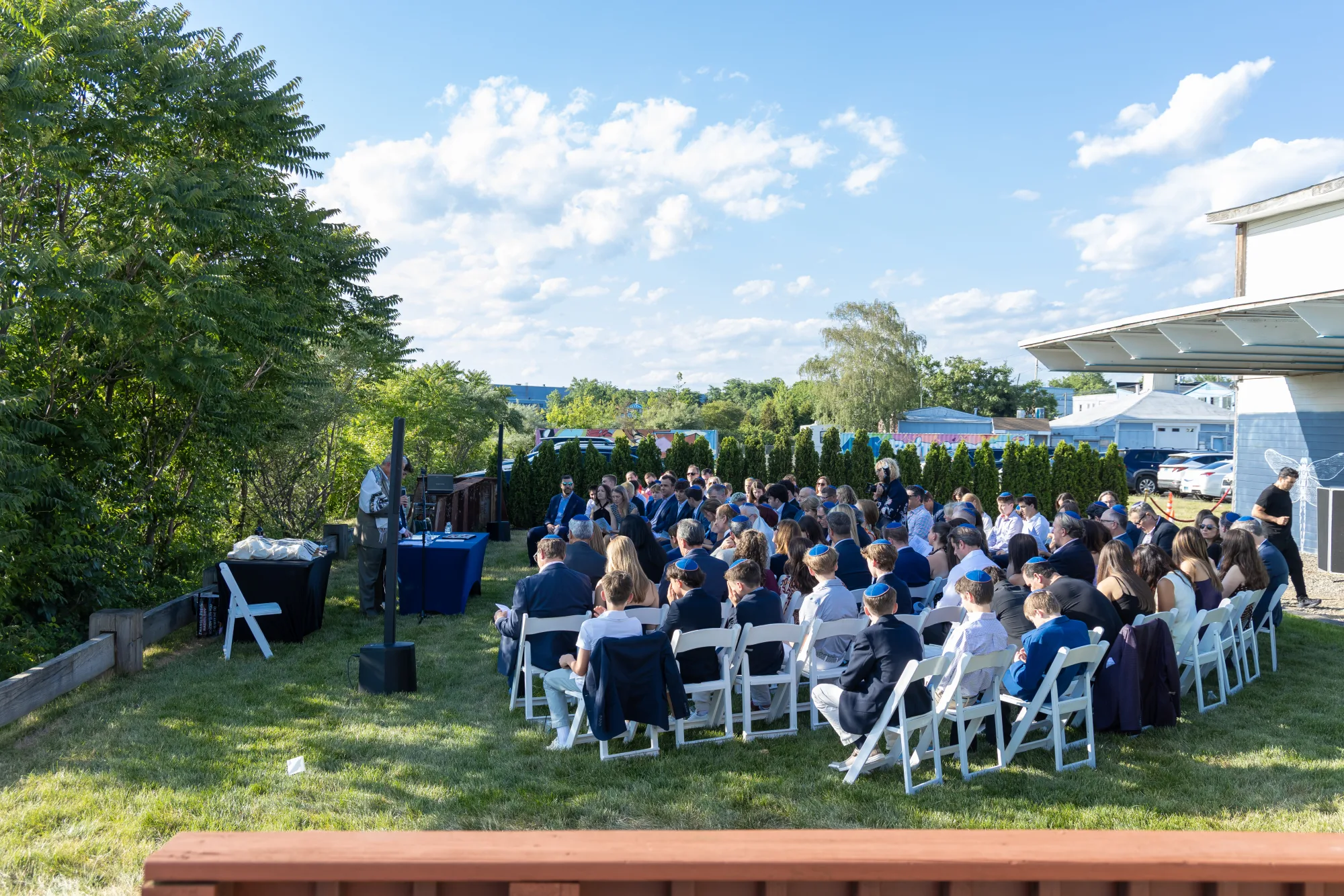 A bar mitzvah service on the waterfront lawn, guests seated in white chairs facing the rabbi, with the mural park walls behind