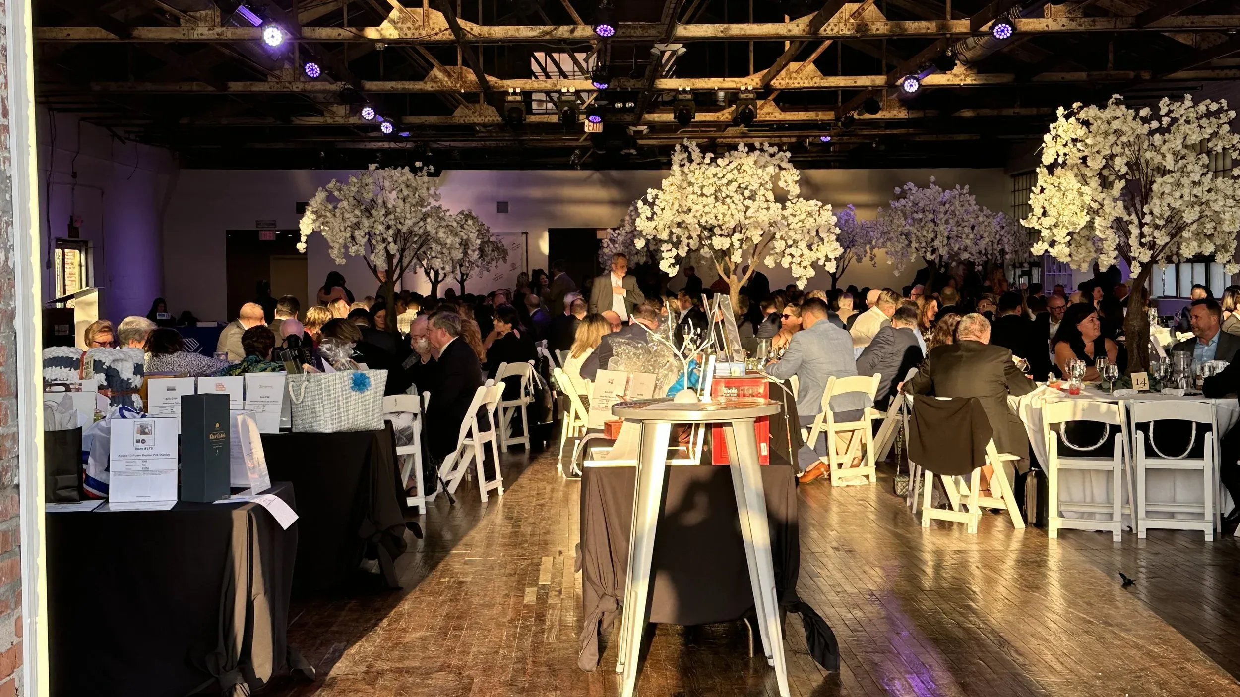 Gala reception with illuminated white cherry-blossom trees under the exposed truss ceiling