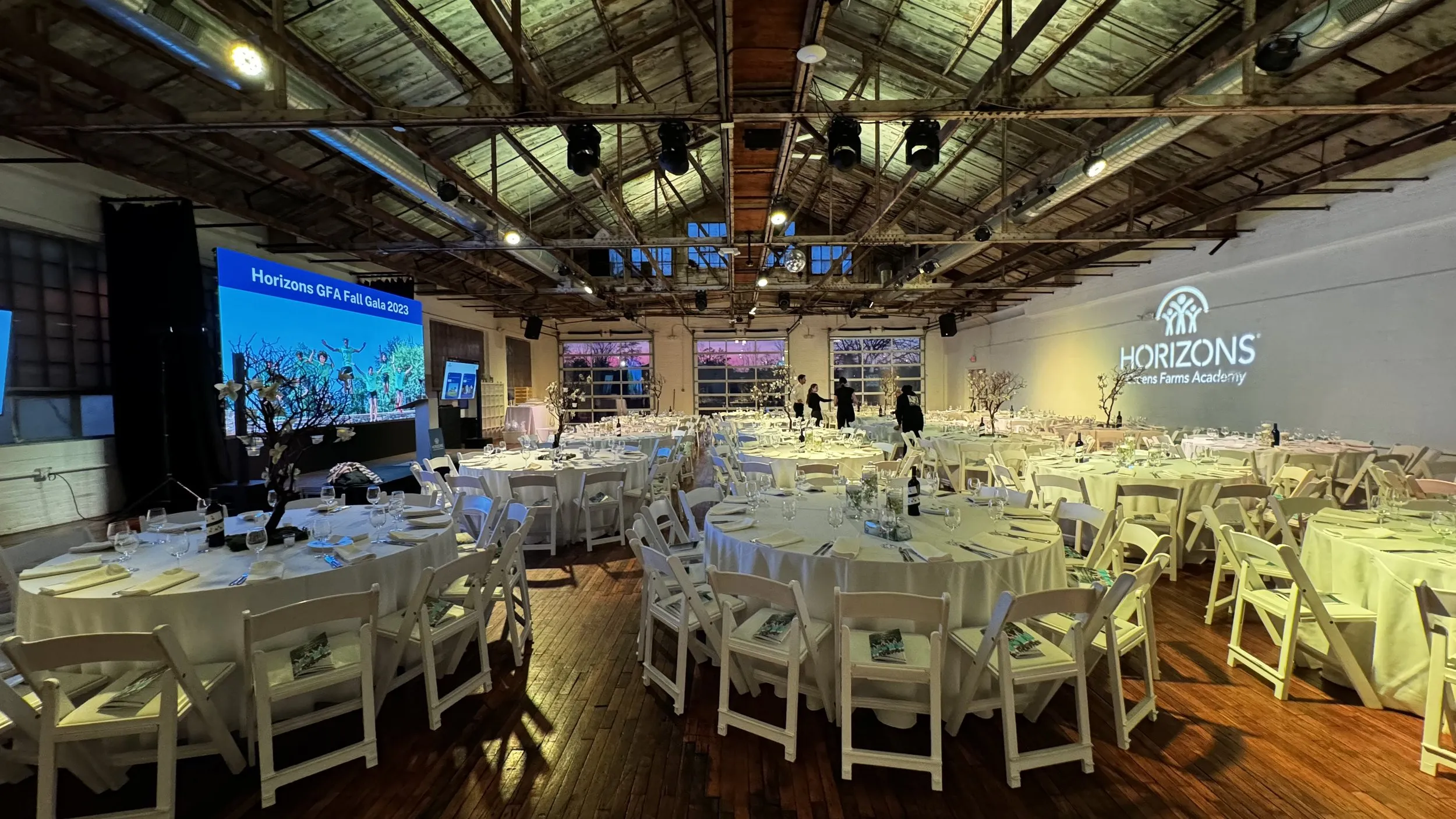 Gala dinner setup under the peaked wood-truss ceiling with garage-door windows at dusk