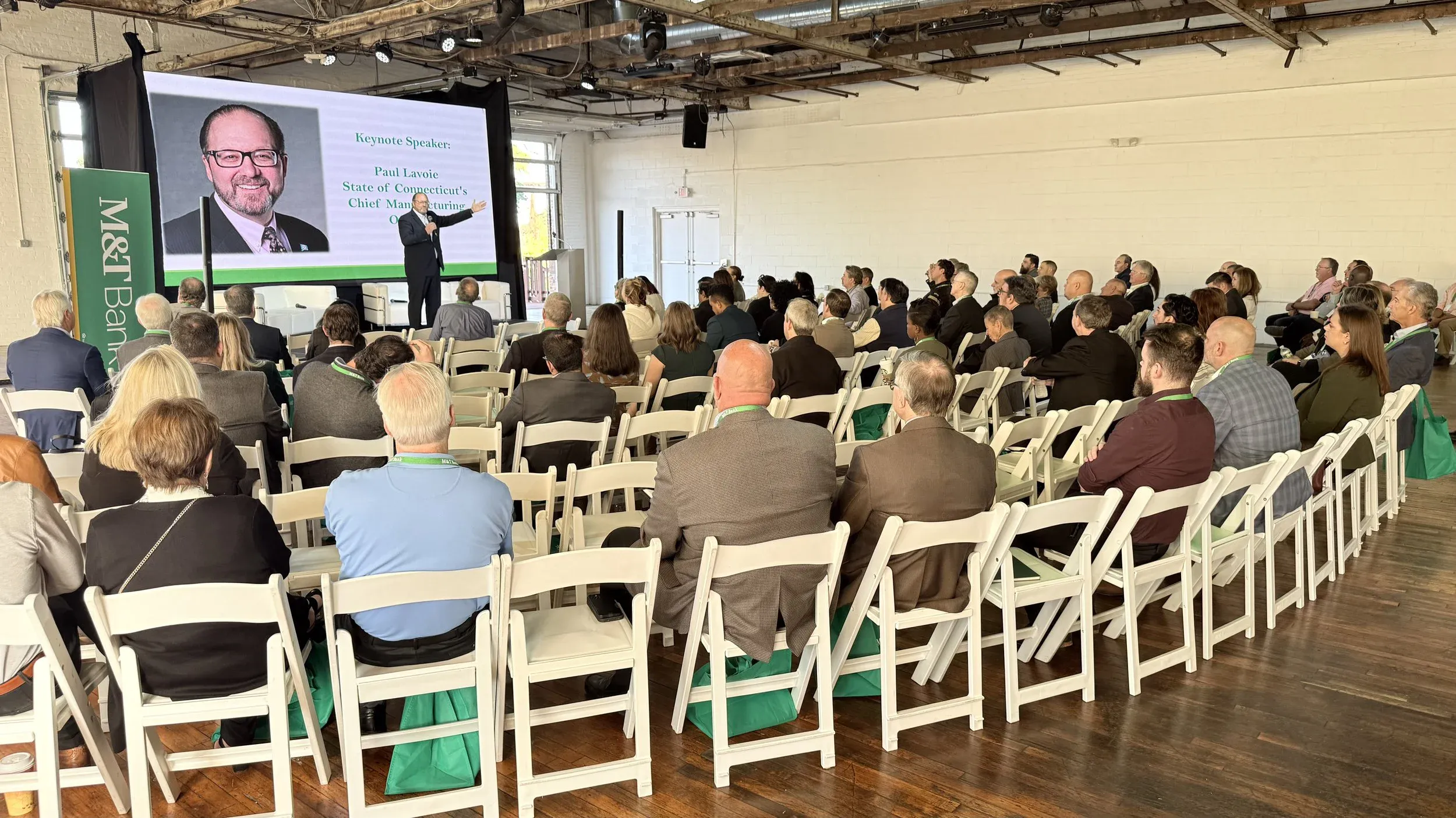 Keynote speaker presenting to a seated audience under the steel-beam ceiling of the Boathouse in daylight