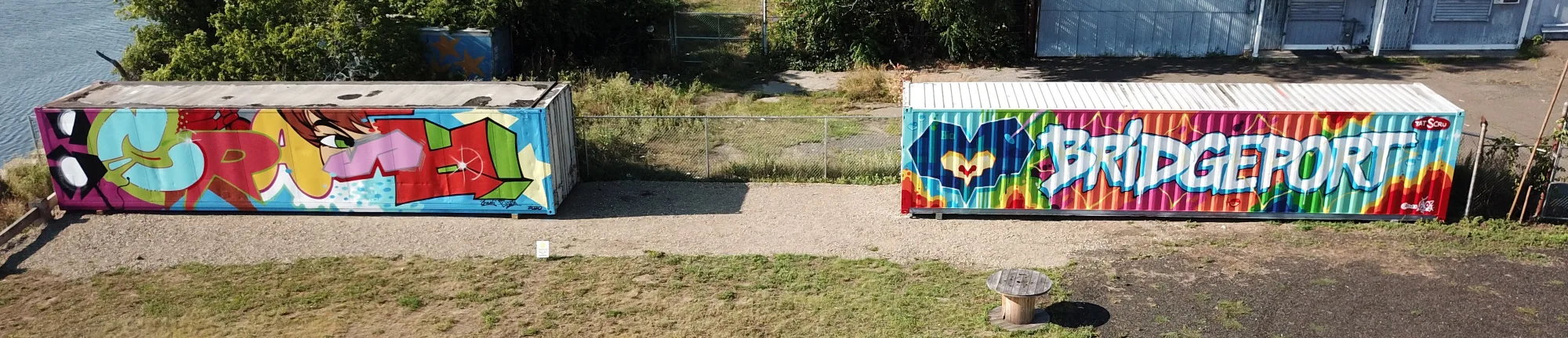 The CrashOne and Tats Cru shipping-container murals side by side on the Pequonnock River waterfront at The Knowlton in Bridgeport, CT.